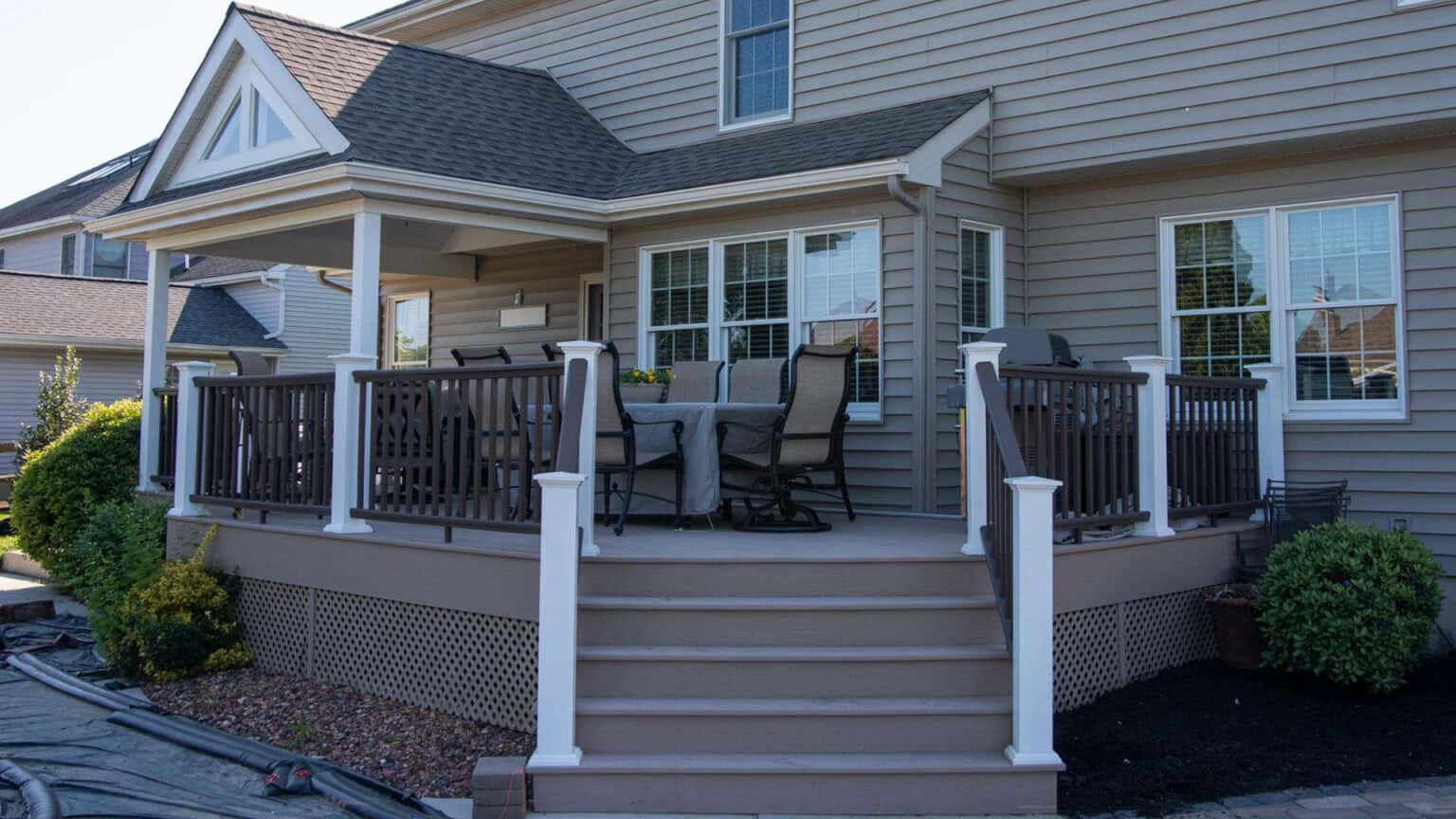 a raised wooden deck attached to a house with beige siding, featuring a dining table and chairs, white railings with dark spindles, and steps leading up to the deck surrounded by landscaped bushes and mulch.