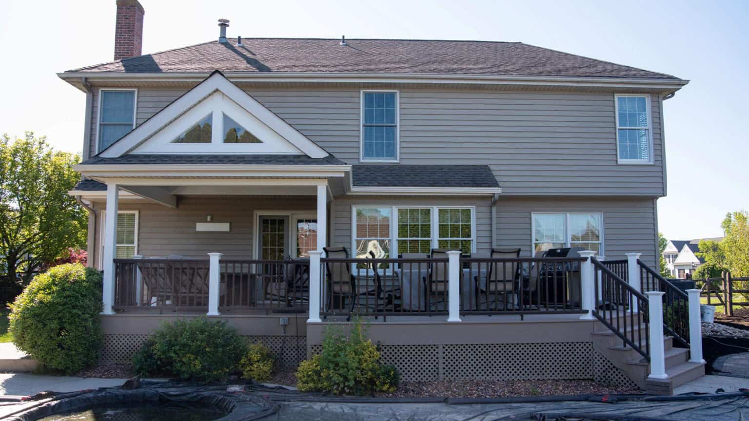 a two story beige house with white trim features a covered back porch and a raised deck with outdoor furniture, railing, and steps leading down to a yard with shrubs and landscaping.