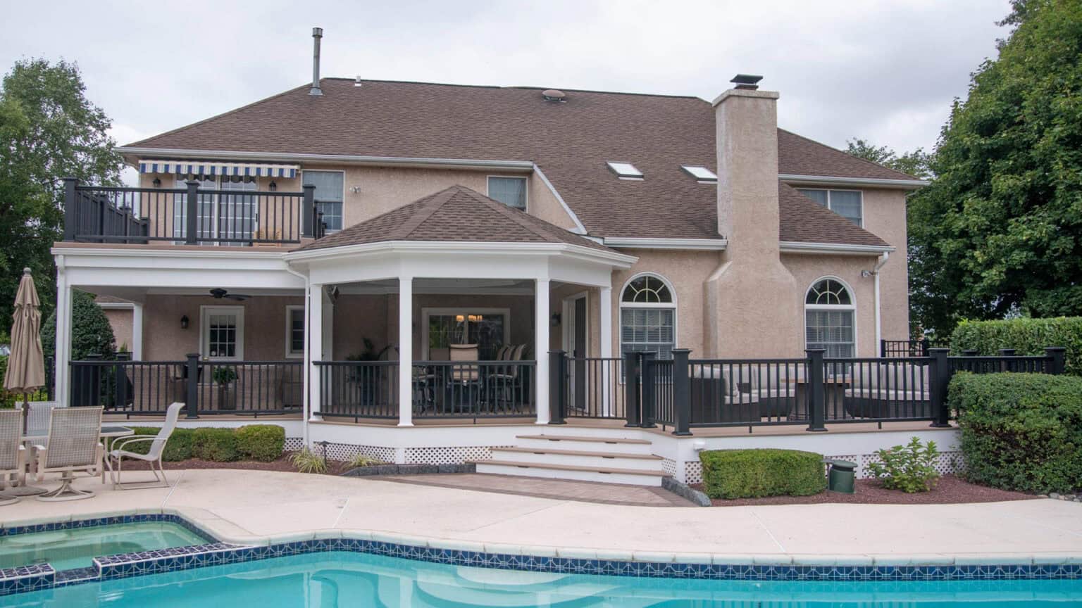 two story beige house with a brown roof, large balcony, and covered patio overlooking a backyard pool. there are black railings, outdoor furniture, and well maintained landscaping.