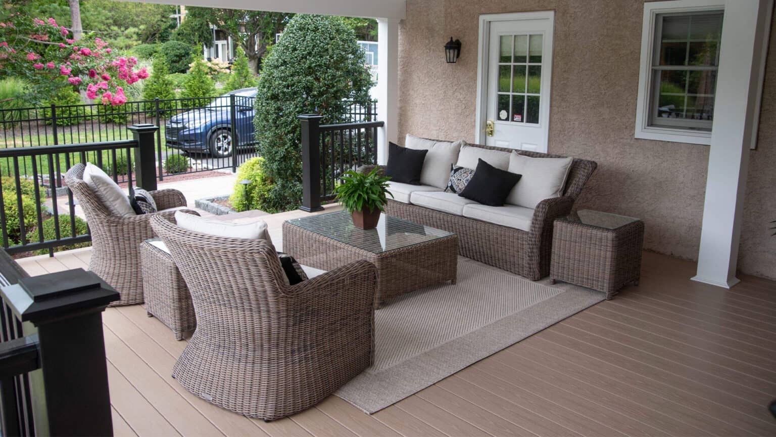 a cozy covered patio with wicker seating, neutral cushions, a glass coffee table, and a potted plant on a beige rug. the area overlooks a green garden and a black car parked in the driveway.