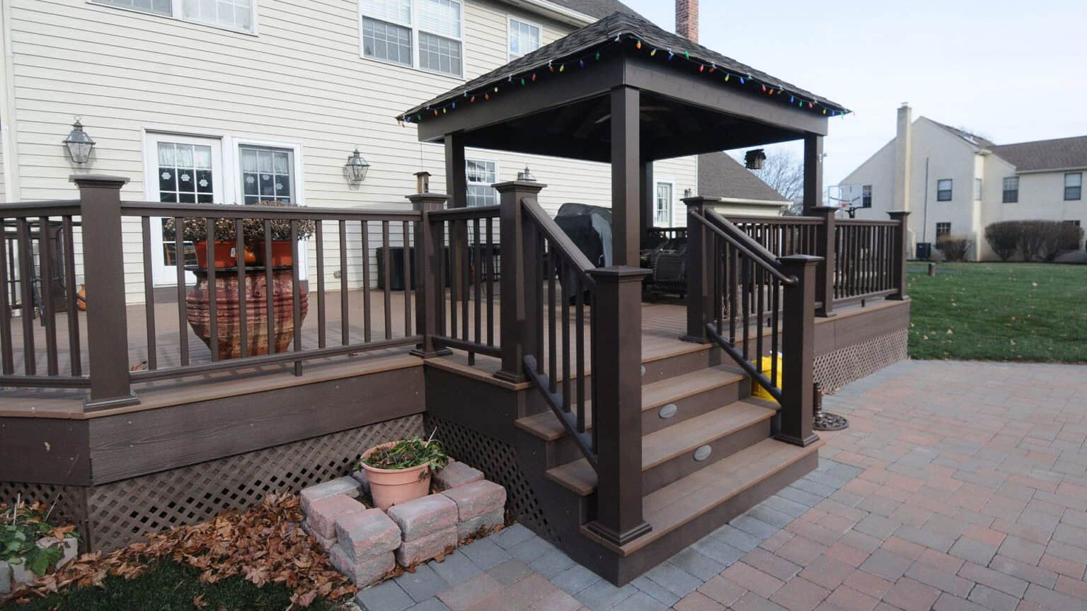 a raised wooden deck with dark railings and steps leads to a covered gazebo. the deck is attached to a beige house with multiple windows. potted plants and a brick patio are in the foreground.