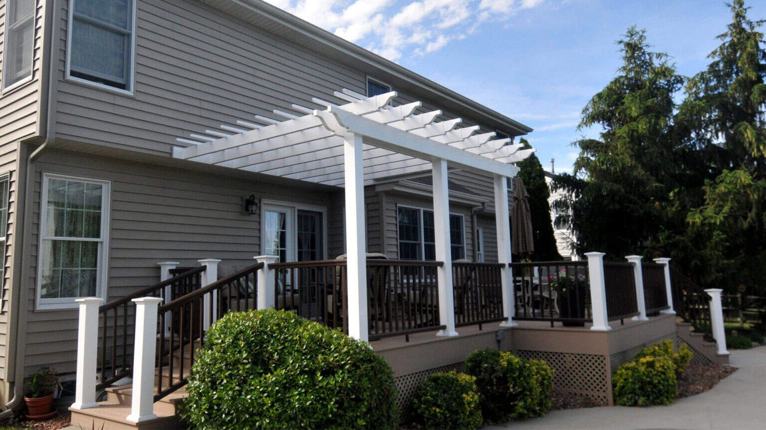 a beige house with a raised wooden deck featuring white railings and a white pergola. green shrubs and small yellow flowers border the deck, and tall trees are visible in the background under a partly cloudy sky.