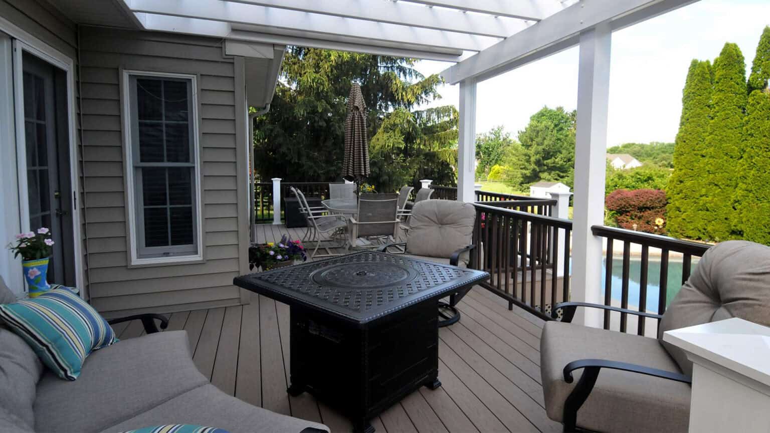 a cozy outdoor patio with cushioned seating, a fire pit table, potted plants, and a dining set under a pergola, overlooking a green yard and swimming pool.
