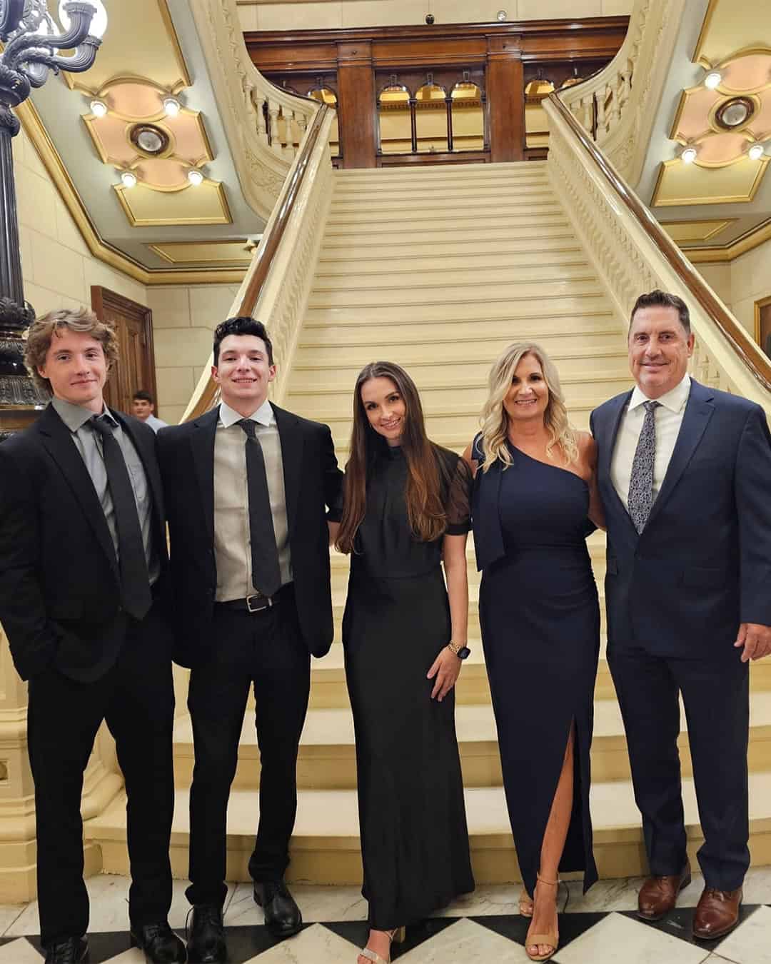 five people dressed formally stand together in front of a grand staircase with ornate lighting, marble floors, and decorative railings in an elegant indoor setting.