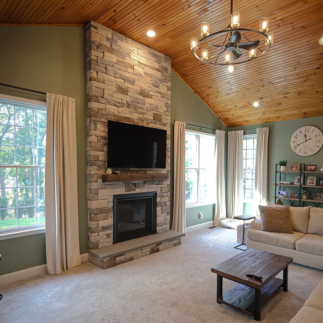 Living room with a floor-to-ceiling stone fireplace, a wall-mounted television, a wood-paneled vaulted ceiling, and a large analog wall clock. Living room with a floor-to-ceiling stone fireplace, a wall-mounted television, a wood-paneled vaulted ceiling, and a large analog wall clock.