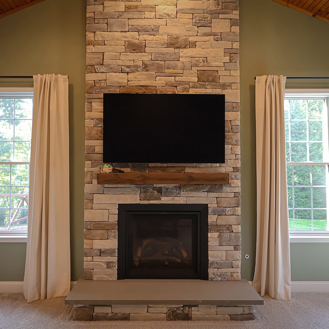A flat-screen television mounted above a stone fireplace with a wooden mantel and stone hearth, flanked by two windows with light-colored curtains. A flat-screen television mounted above a stone fireplace with a wooden mantel and stone hearth, flanked by two windows with light-colored curtains.
