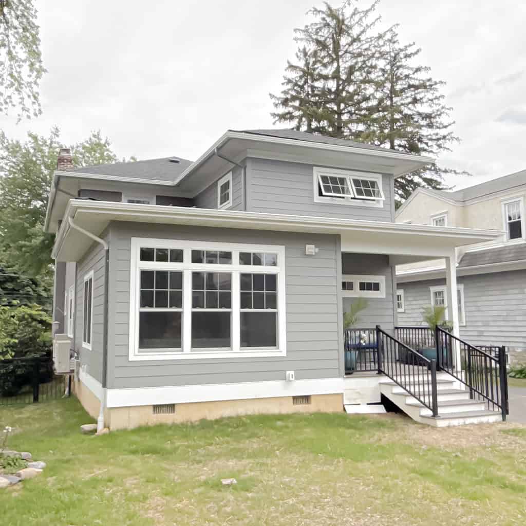 a modern two story gray house with white trim, large windows, and a small porch with black railings. the yard is partially grass covered, and tall trees are visible in the background under a cloudy sky.