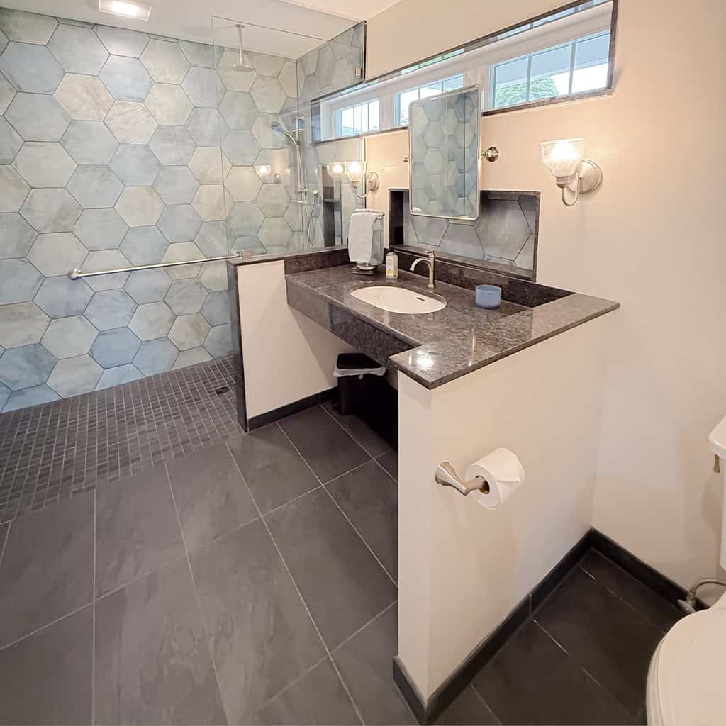 modern bathroom with large hexagonal blue tiles in the shower, a dark stone countertop with undermount sink, wall mounted lights, a wide mirror, gray floor tiles, and a toilet partially visible.