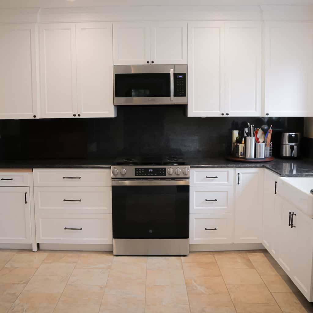 a modern kitchen with white cabinets, black countertops, a stainless steel oven and microwave, and various kitchen utensils and appliances in the corner near a sink. light tile flooring is visible.
