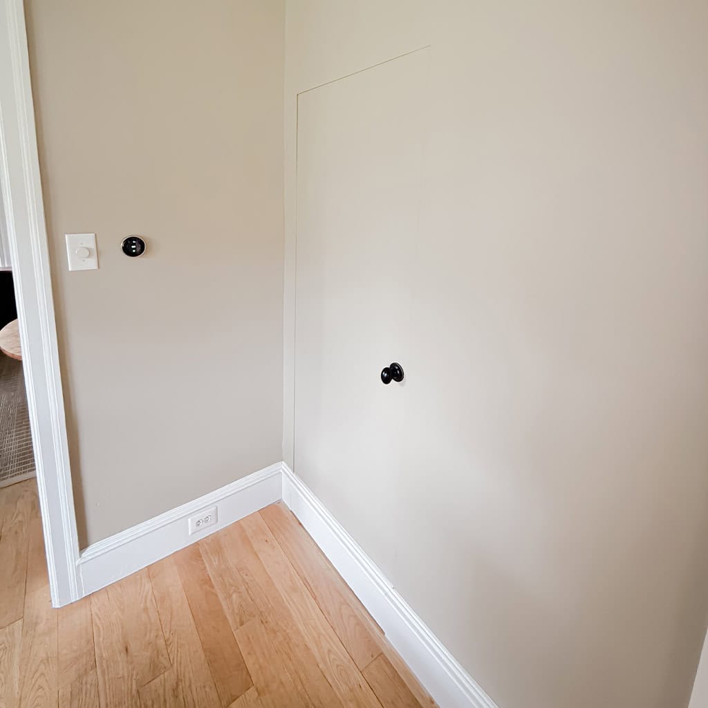 a small, closed door with a black knob is set into a beige wall above light wood flooring. there is a thermostat and a light switch on the wall to the left of the door.