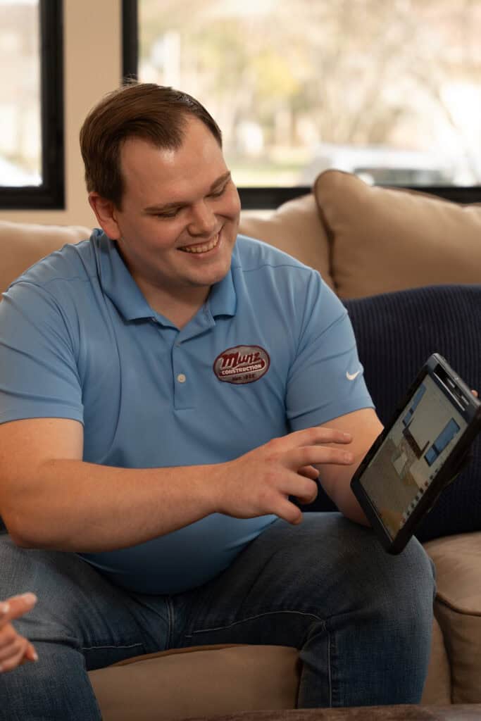 a smiling man in a light blue shirt sits on a beige couch and points at a tablet, which displays interior design images.