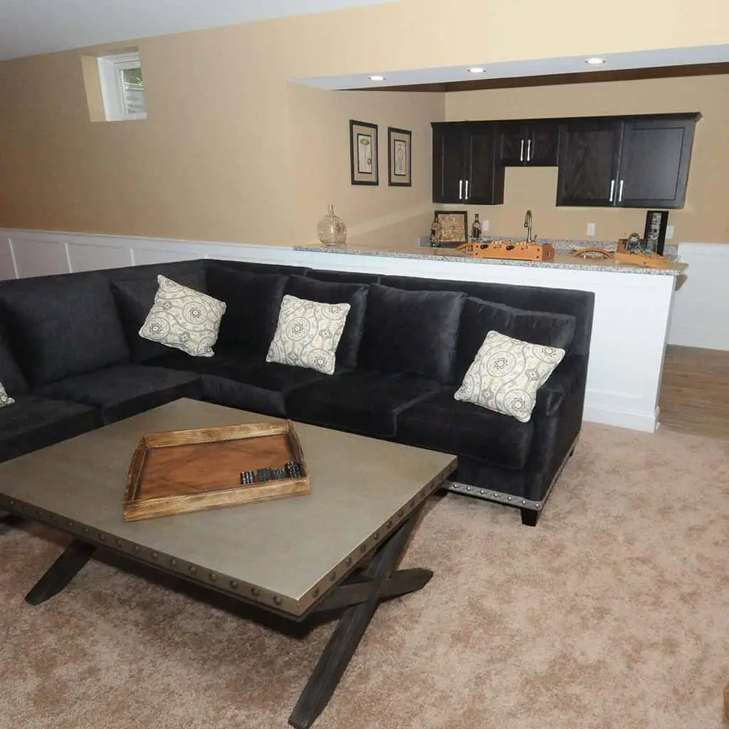a cozy living room with a dark gray sectional sofa, patterned pillows, a wooden coffee table with a tray, and a small kitchen area with dark cabinets in the background. the carpet is light brown.