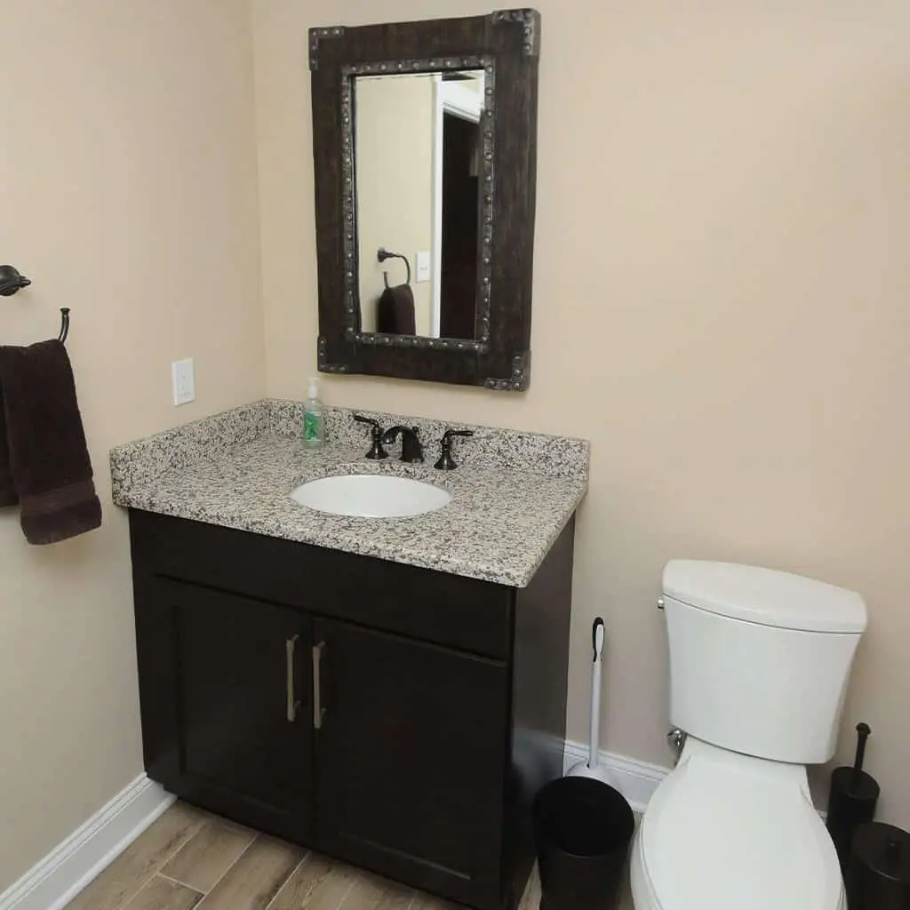 a bathroom with a granite countertop vanity, dark cabinets, an oval sink, a wall mounted mirror with a dark frame, a white toilet, a black trash can, and beige walls with wood look tile flooring.
