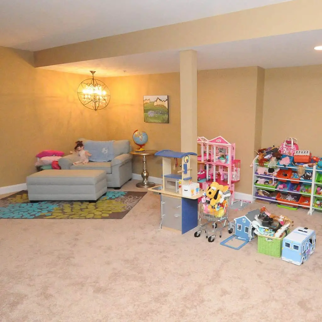 a playroom with tan walls and carpet, a gray sectional sofa with a doll, a globe on a side table, wall art, and shelves filled with toy houses, stuffed animals, and various playthings scattered on the floor.