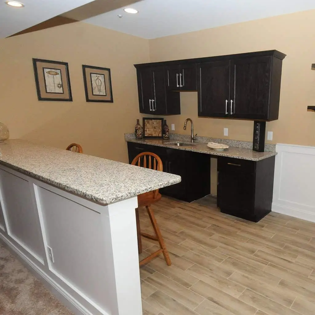a small home bar area with granite countertops, dark wood cabinets, a sink, two wooden barstools, framed artwork on a tan wall, and light wood style tile flooring.