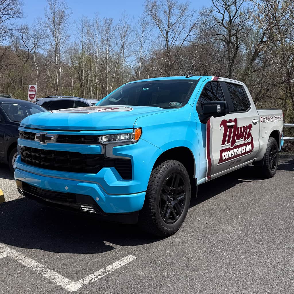 a blue and white chevrolet pickup truck with “munz construction” branding is parked in a lot on a sunny day, with trees and other vehicles in the background.