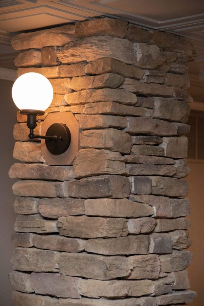 a round wall sconce with a white globe bulb is mounted on a pillar made of stacked, uneven stone blocks in an indoor setting.