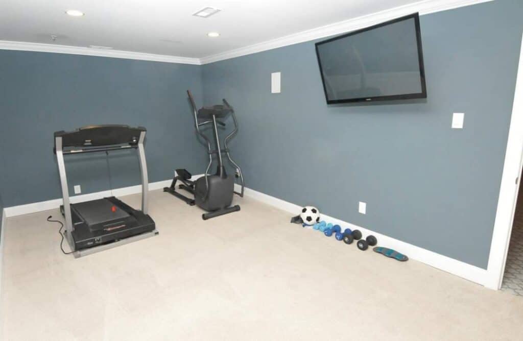 a home gym with a treadmill, elliptical machine, wall mounted tv, and a row of small dumbbells, a jump rope, a yoga mat, and a soccer ball on a carpeted floor against blue walls.