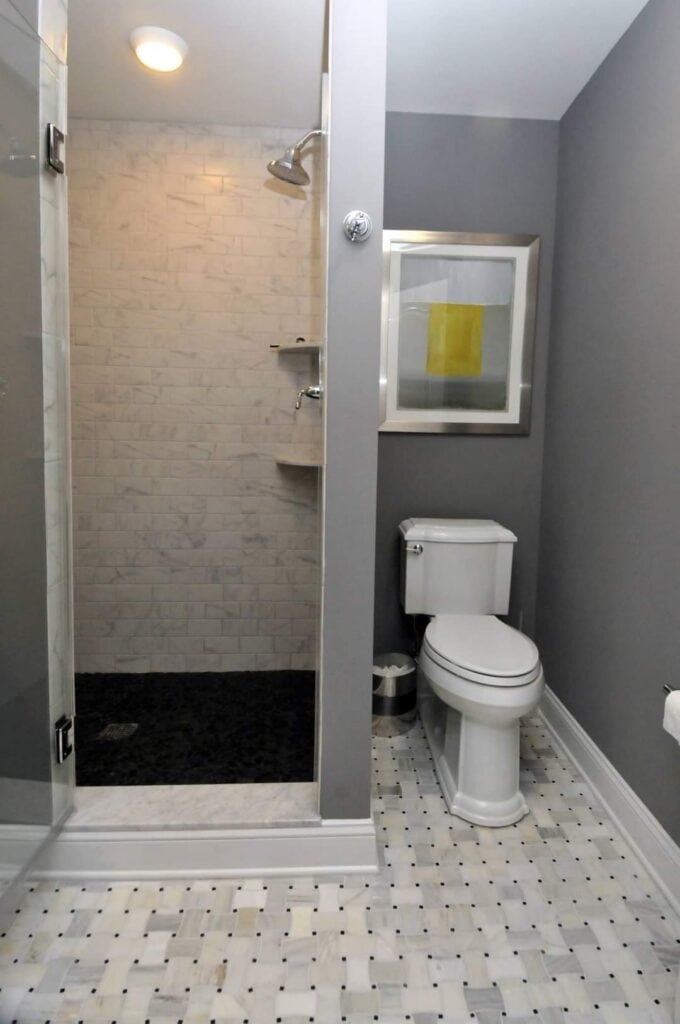 a modern bathroom with gray walls, a white toilet, a small waste bin, a walk in shower with white tile, a glass door, and a framed abstract artwork above the toilet. the floor has a white and gray mosaic tile pattern.