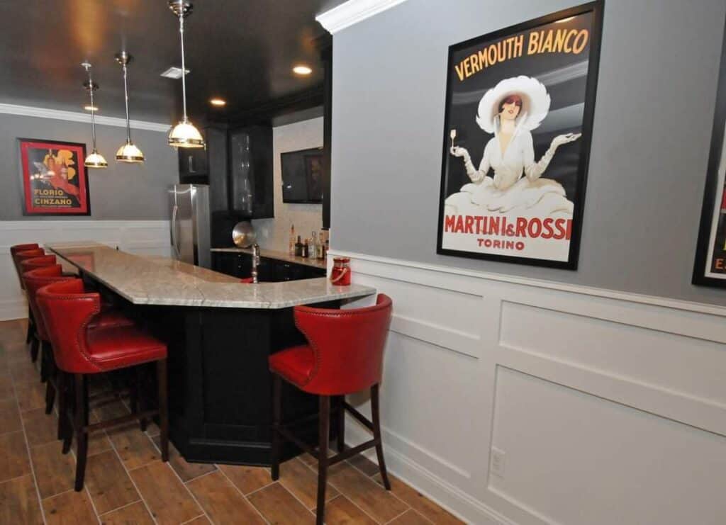 a home bar area with a marble countertop, six red upholstered bar stools, pendant lights above, wood look floor, black cabinetry, and vintage posters on gray and white paneled walls.