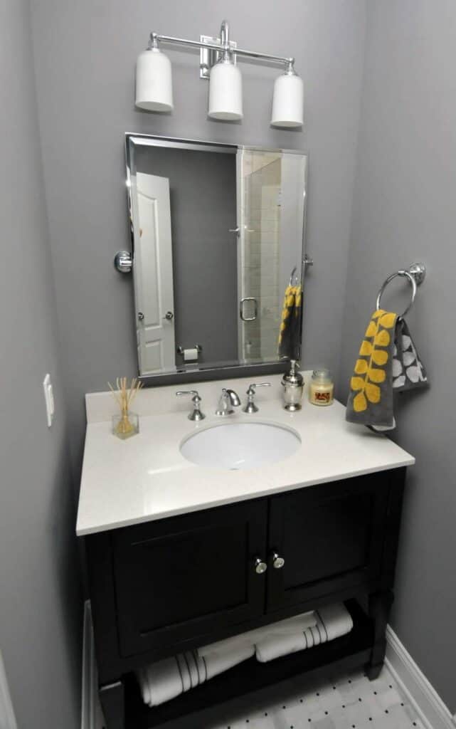 modern bathroom with light gray walls, a white countertop sink, dark vanity cabinet, and rectangular mirror. above the mirror are three lights. hand towel with yellow leaf pattern hangs on the right. towels are stored below the sink.