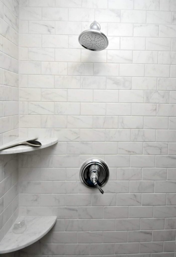 a modern shower with white subway tile walls, a silver showerhead, a round control handle, and two corner shelves&mdash;one holding a silver razor.