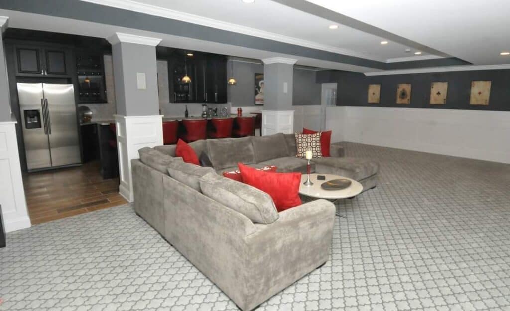 a spacious basement lounge with a large gray sectional sofa, red accent pillows, a round coffee table, and a kitchenette featuring black cabinets and stainless steel appliances. the floor is carpeted with a light geometric pattern.