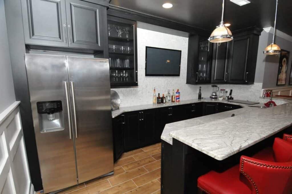 modern kitchen with dark cabinets, stainless steel refrigerator, mounted tv, glass shelves with glassware, marble countertop, wooden floor, and red bar stools along a curved breakfast bar.