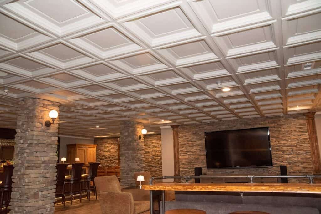 a cozy bar lounge with stone pillars, a coffered ceiling, a large wall mounted tv, wooden bar stools, and a stone accent wall, warmly lit by wall sconces and table lamps.