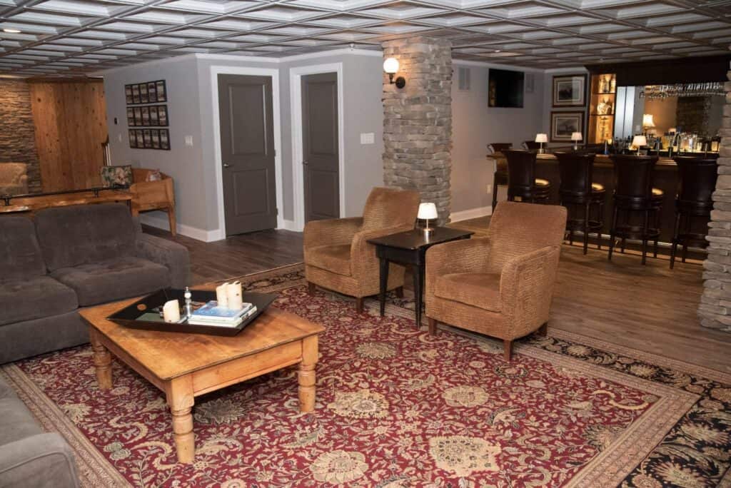 a cozy basement living area with a patterned rug, brown sofa, wooden coffee table, two wicker chairs, and a bar area with high stools. stone pillars and soft lighting create a warm, inviting atmosphere.