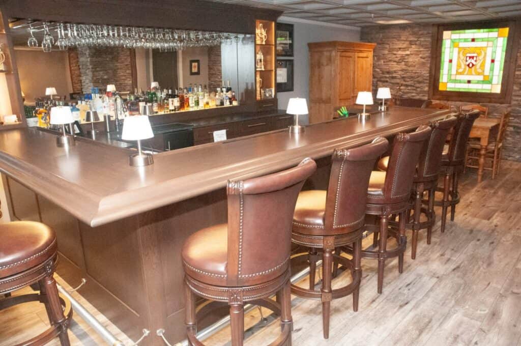 a polished wooden bar with brown leather barstools, bottles of liquor behind the counter, small lamps on the bar, and a stained glass window on the wall in a cozy, well lit room with wood and stone accents.
