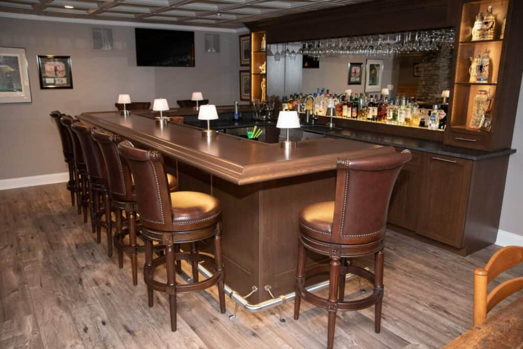 a home bar with brown leather barstools, a wooden counter, a mirrored back wall, shelves stocked with bottles, glasses hanging overhead, and framed art on gray walls. the floor is wood, and the space is warmly lit.