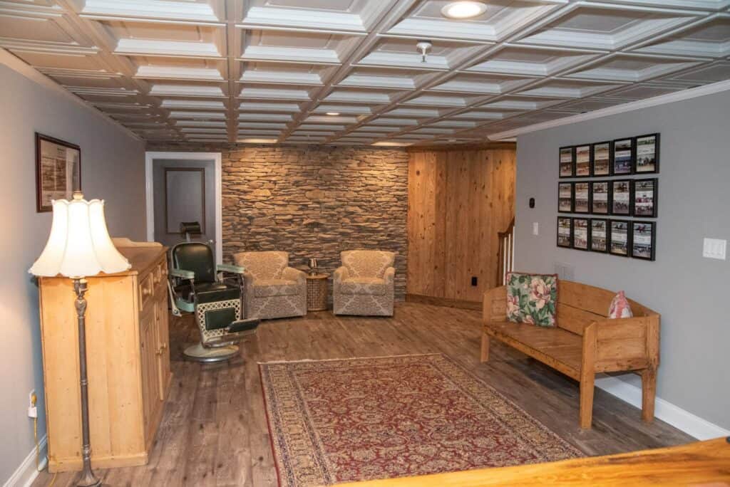 a cozy basement room with a patterned ceiling, stone accent wall, two armchairs, vintage barber chair, wooden bench with floral pillow, framed photos on the wall, area rug, and a wooden lamp on a small bar.