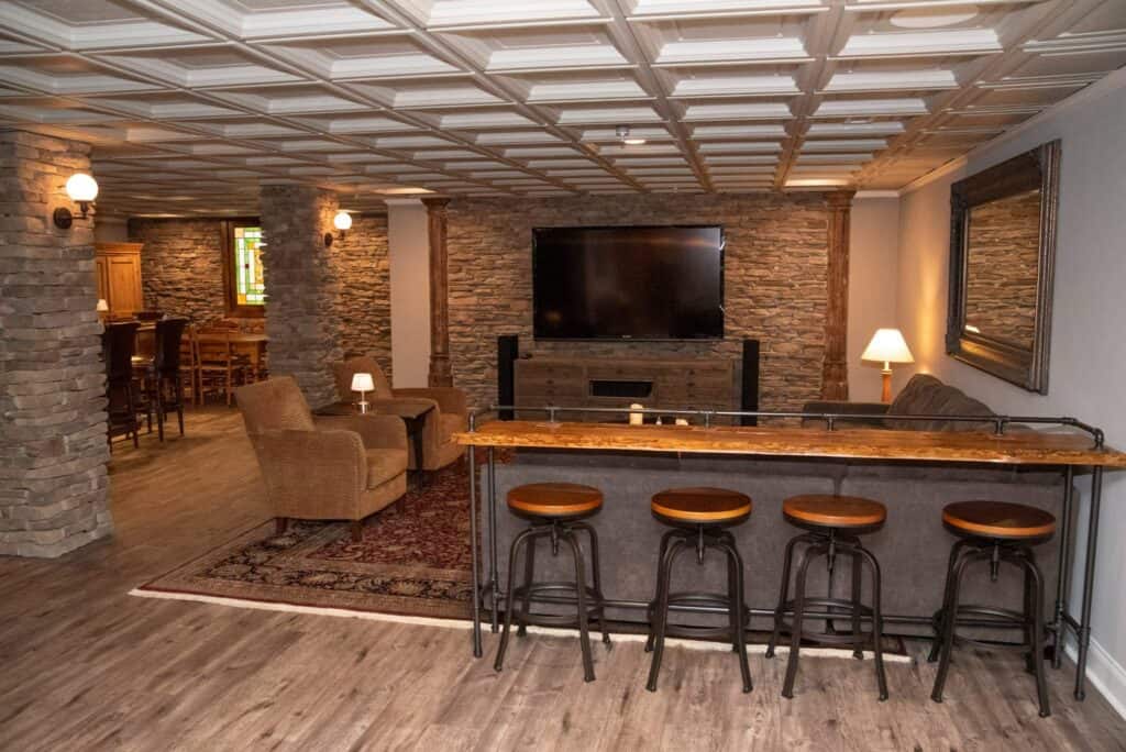 a cozy basement lounge with stone walls, a large tv, fireplace, wicker armchairs, a bar with four stools, a wooden table, lamps, and a decorative ceiling. a dining area is visible in the background.