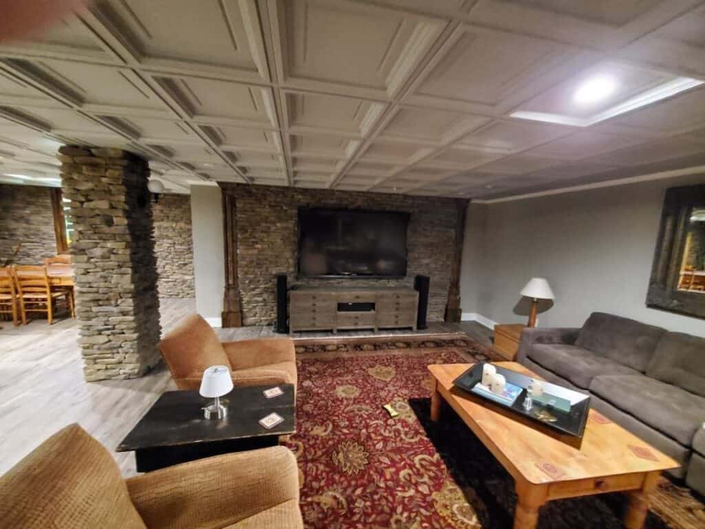 a cozy living room with stone walls, a large wall mounted tv, brown armchairs, a gray couch, a red patterned rug, and a wooden coffee table with decor. a dining area is visible in the background.