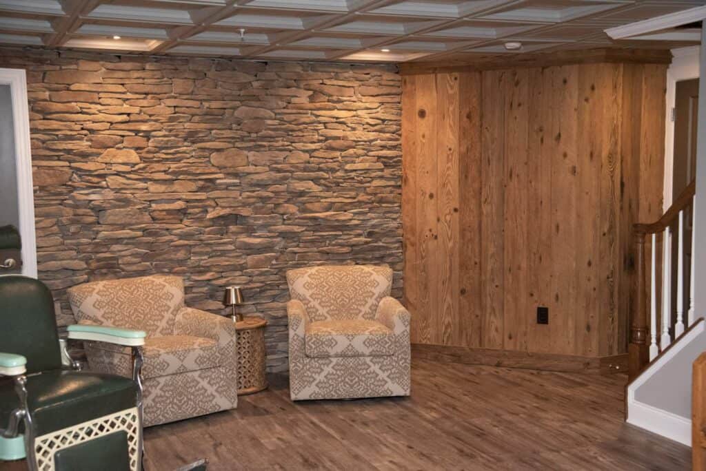 two patterned armchairs sit beside a small side table on wood style flooring; the background is a mix of rustic stone and wooden walls, with a staircase visible on the right.