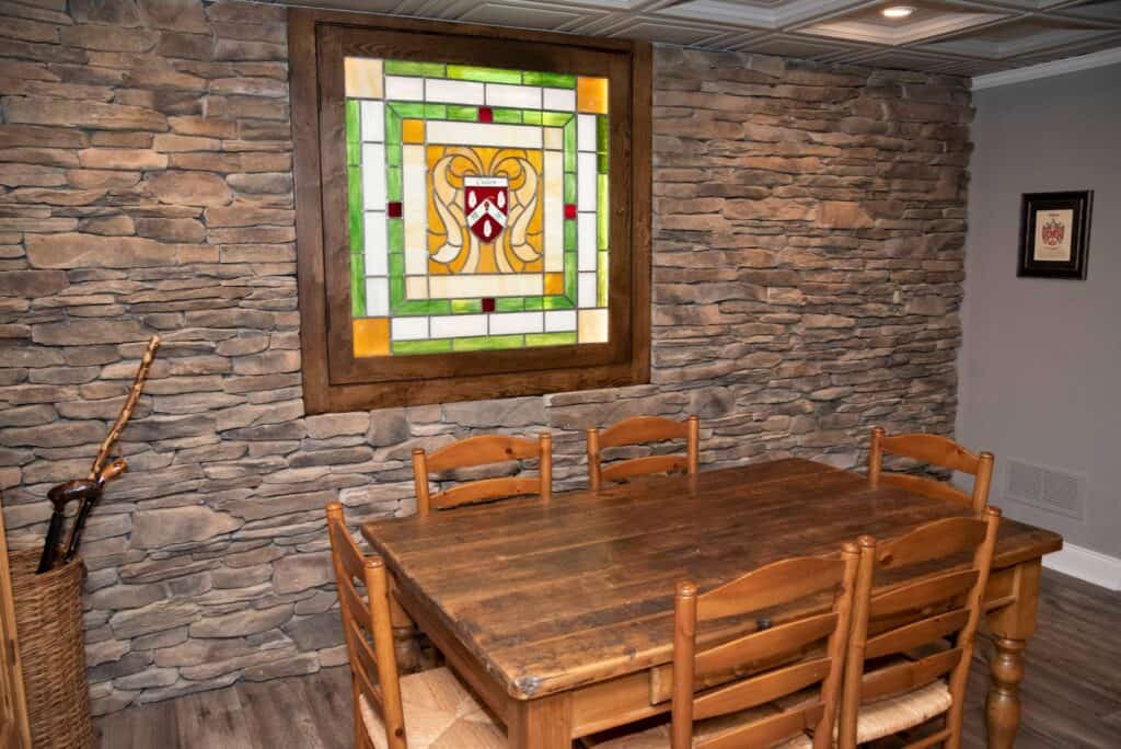 a rustic dining room with a wooden table and six chairs, stone accent wall, and a large stained glass panel with a crest design. a basket with a sword in the corner and framed art on a side wall.