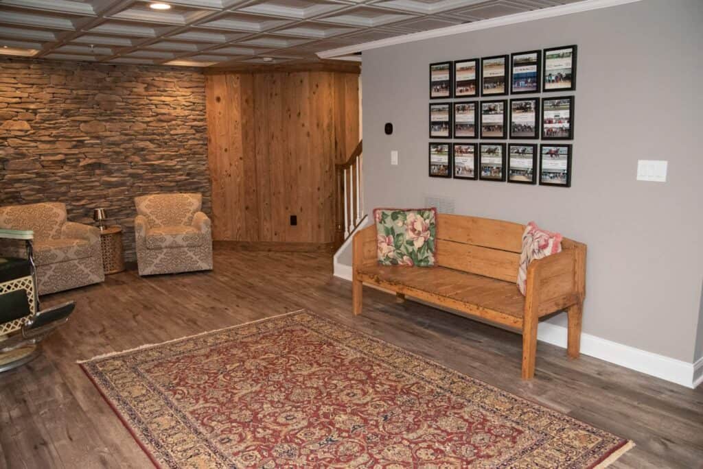 a cozy room with wooden floors, a patterned area rug, a wooden bench with floral pillows, two upholstered chairs, a stone accent wall, and a photo collage on the wall near a wooden staircase.
