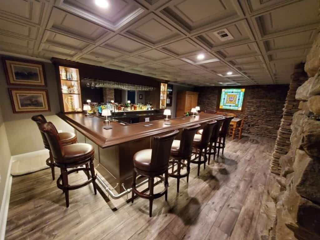 a cozy home bar with a wooden counter, high backed chairs, shelves with bottles and glasses, a stained glass window, stone walls, wood floors, and a small dining area in the corner.