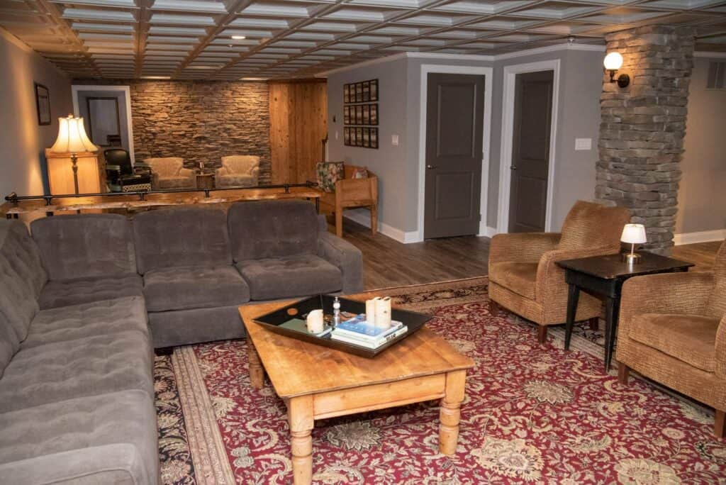 a cozy living room with a gray sectional sofa, two wicker armchairs, a wooden coffee table on a patterned red rug, stone accent walls, and warm lighting, creating a comfortable and inviting atmosphere.