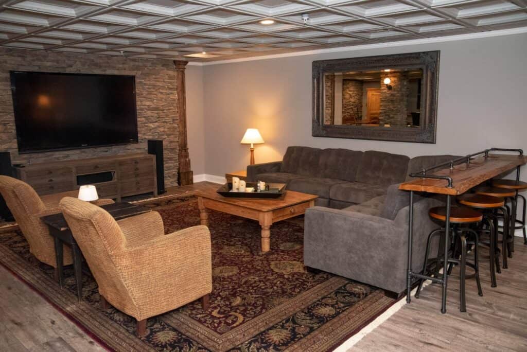 a cozy living room with a large tv, stone accent wall, gray sectional sofa, two wicker armchairs, a wooden coffee table, side lamp, large wall mirror, patterned rug, and a high wooden bar with stools.