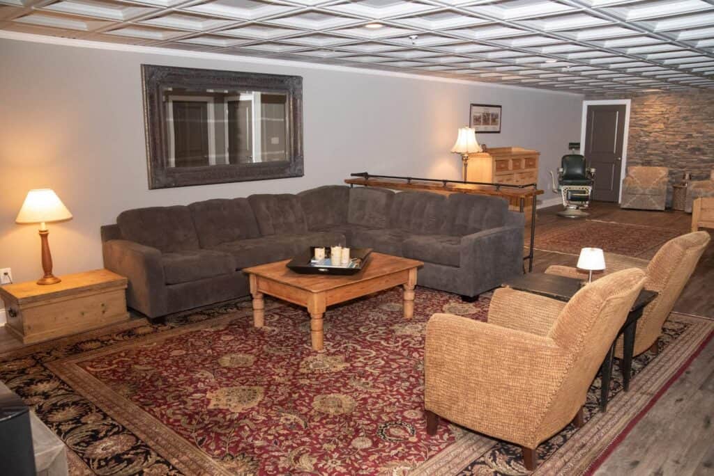 a cozy living room with a large gray sectional sofa, two wicker armchairs, a wooden coffee table, side tables with lamps, a red patterned rug, and neutral decor, including a large mirror on the wall.