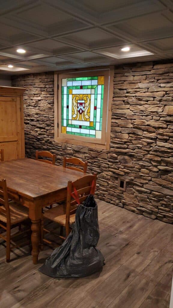 a rustic dining room with a wooden table and chairs, stone textured walls, a stained glass window, and a large black trash bag on the wooden floor.