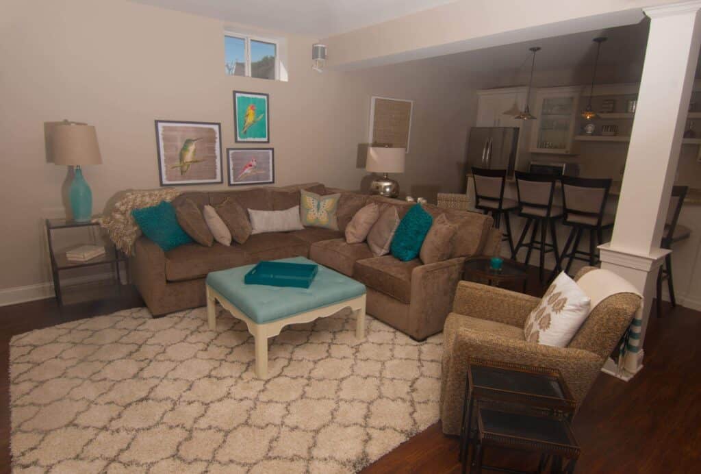 a cozy living room with a brown sectional sofa, patterned pillows, armchair, cream rug, and light blue coffee table. art and lamps decorate the space. a kitchen with barstools is visible in the background.