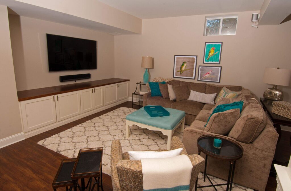 a cozy living room with a large brown sectional sofa, patterned rug, armchair, and teal accents. art with birds hangs on the wall, and a flat screen tv is mounted above a long cabinet. lamps and small tables complete the space.