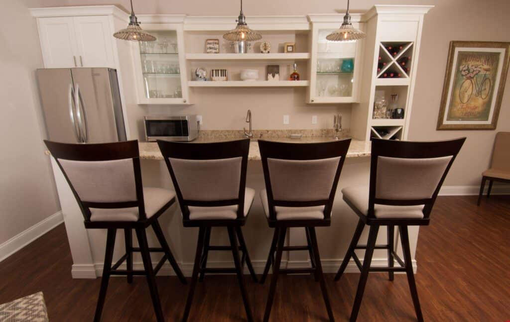 a modern kitchen with four upholstered barstools at a marble counter, pendant lights above, white cabinets, a stainless steel refrigerator, a microwave, and a wine rack. a framed bicycle artwork hangs on the wall.