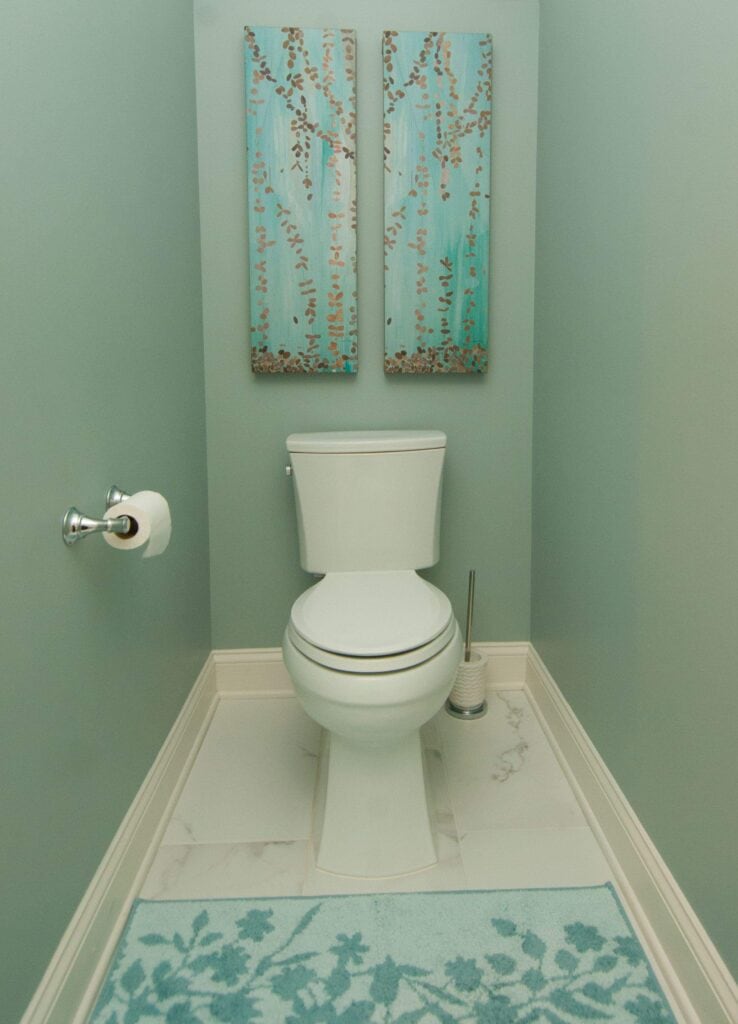 a white toilet with a roll of toilet paper and a toilet brush stands in a narrow bathroom with light blue walls, white tile floor, a blue floral rug, and two blue floral paintings hanging on the wall above the toilet.