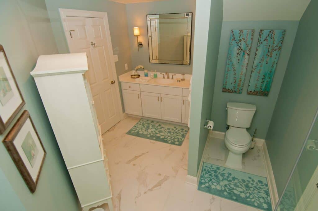 a modern bathroom with teal walls, white cabinetry, a large mirror above the sink, white toilet, artwork on the walls, and teal patterned rugs on a light marble floor.