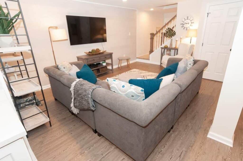 a cozy living room with a large gray sectional sofa, blue and patterned pillows, a tv mounted on the wall, a wooden console, floor lamp, and neutral decor on light wood flooring. stairs and a clock are visible in the background.