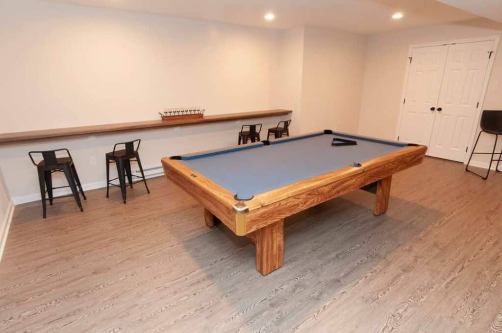 a spacious room with a wooden pool table in the center, four black metal chairs along a bar height counter against the wall, and light wood flooring. the room is well lit with recessed ceiling lights.